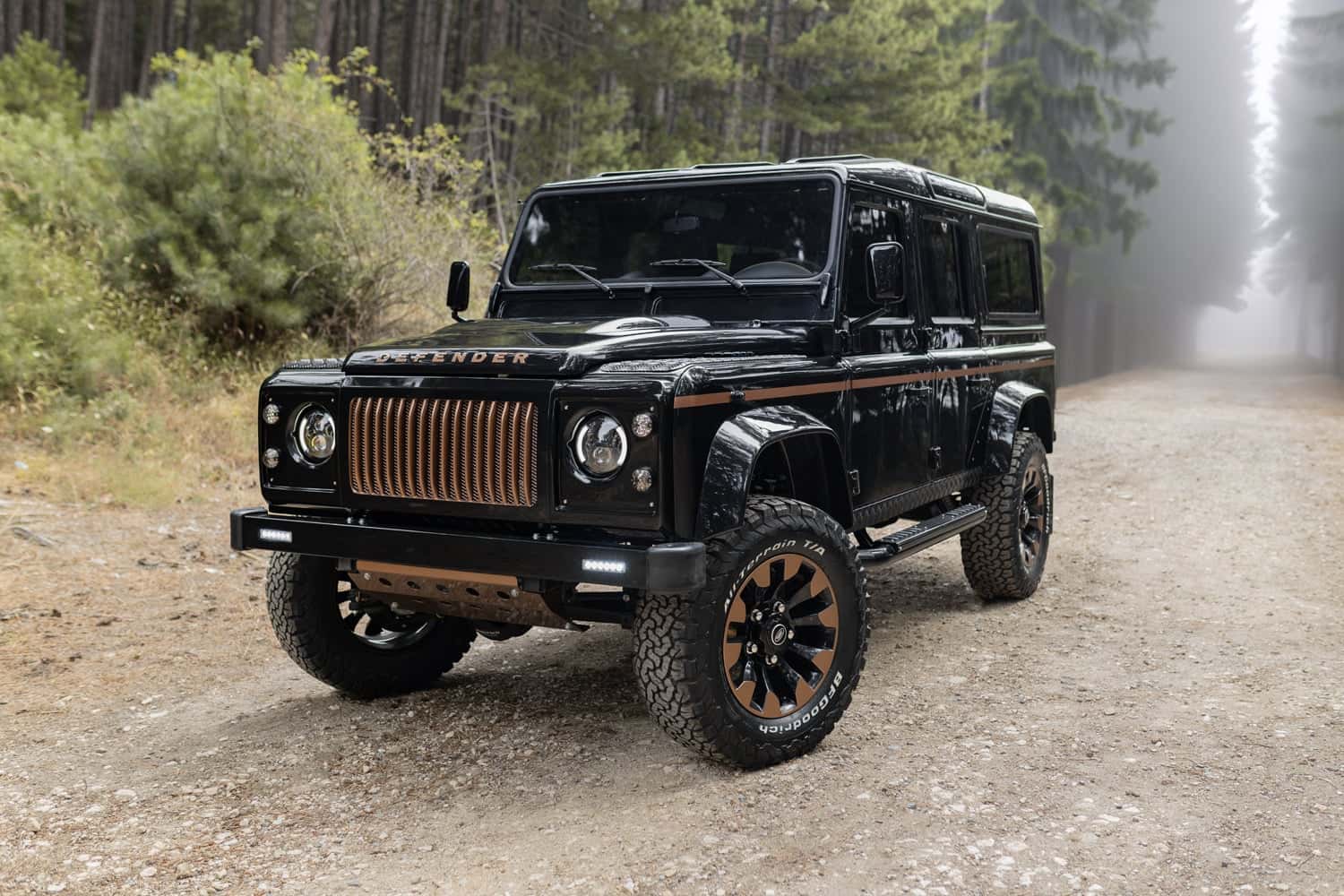 Custom black Land Rover Defender with bronze accents parked on a forest road