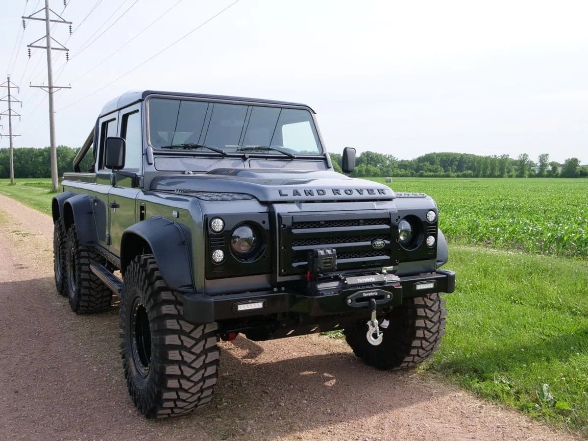 Black modified Land Rover Defender parked on a dirt road with lush green fields in the background