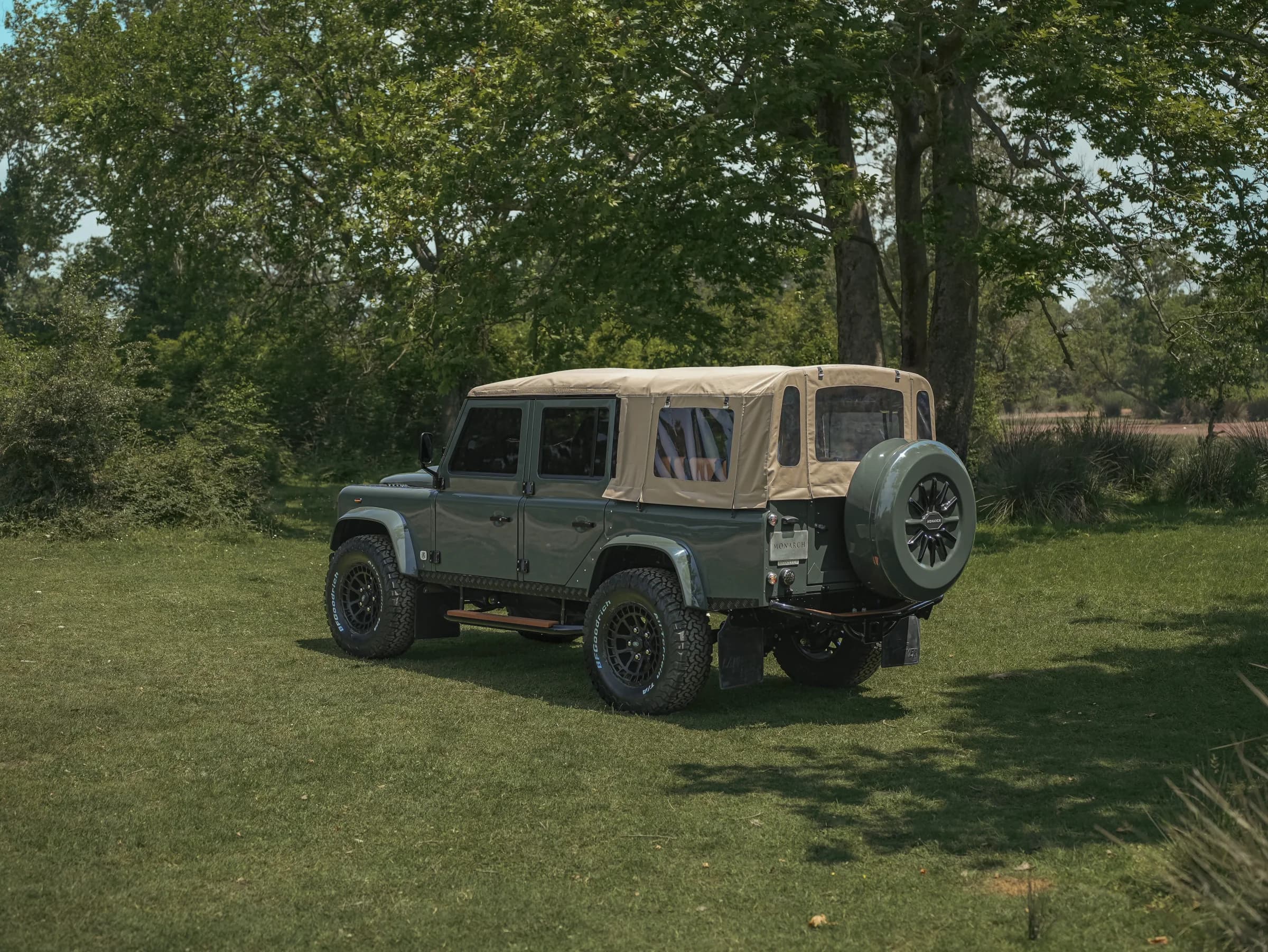A bespoke Land Rover Defender with a tan soft top parked on grass, surrounded by trees.