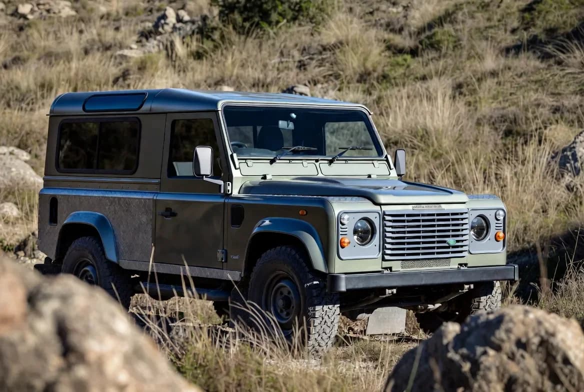 A 1990 Land Rover Defender 90 in profile, showing the classic boxy silhouette with the newly applied Defender badge on the front, parked on rugged terrain
