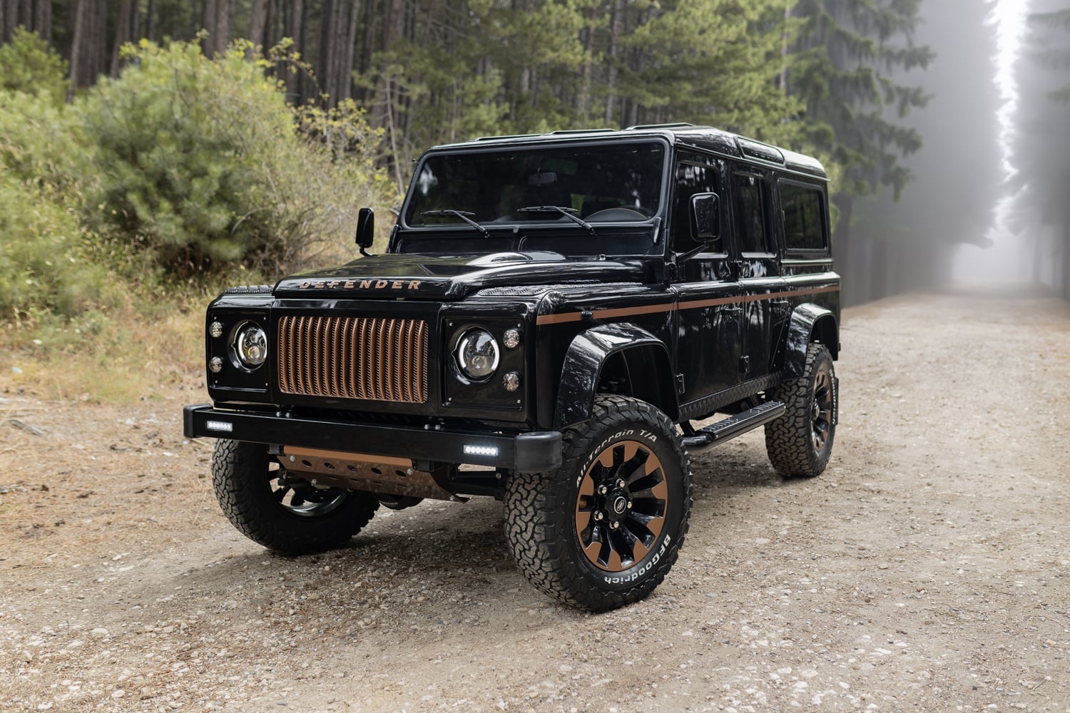 Custom black Land Rover Defender with bronze accents parked on a forest road