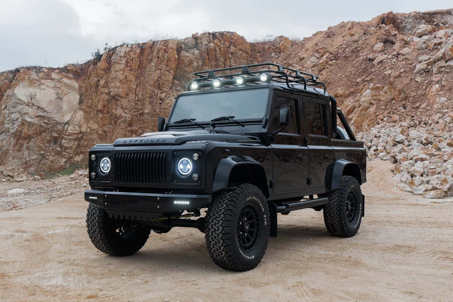 Front View: Custom Land Rover Defender Black custom Land Rover Defender with off-road tires and roof rack in rugged terrain