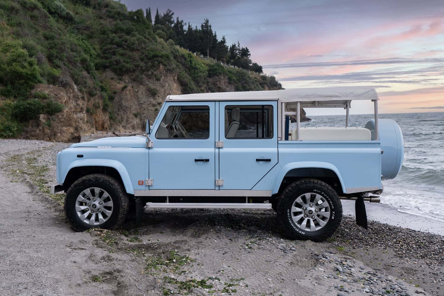 Side View: Bespoke Land Rover Defender A custom Land Rover Defender in light blue parked on a beach with a sandy shoreline and ocean waves.