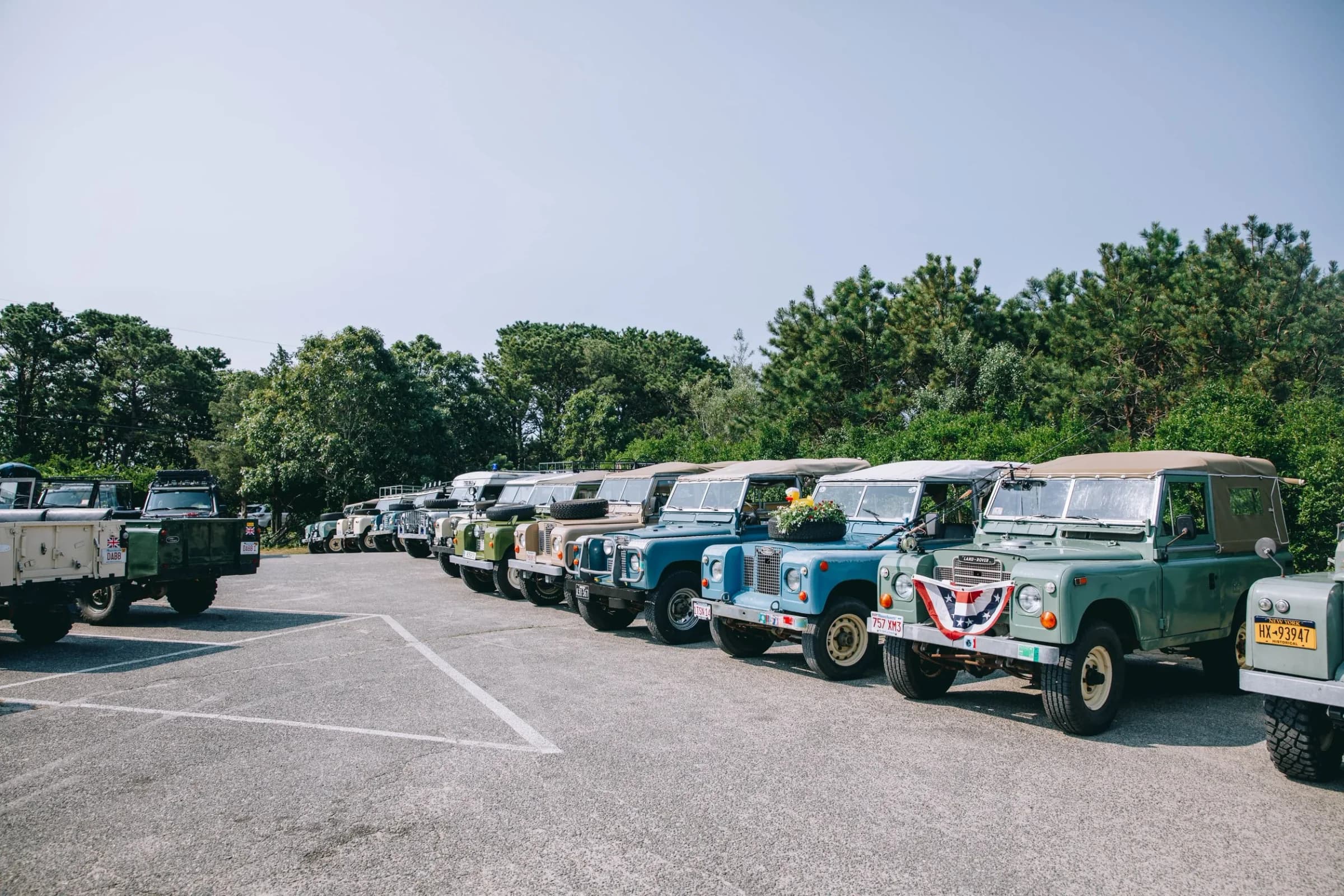 A row of classic Defenders lined up at a coastal community event, bonnets open, owners gathered in small groups talking