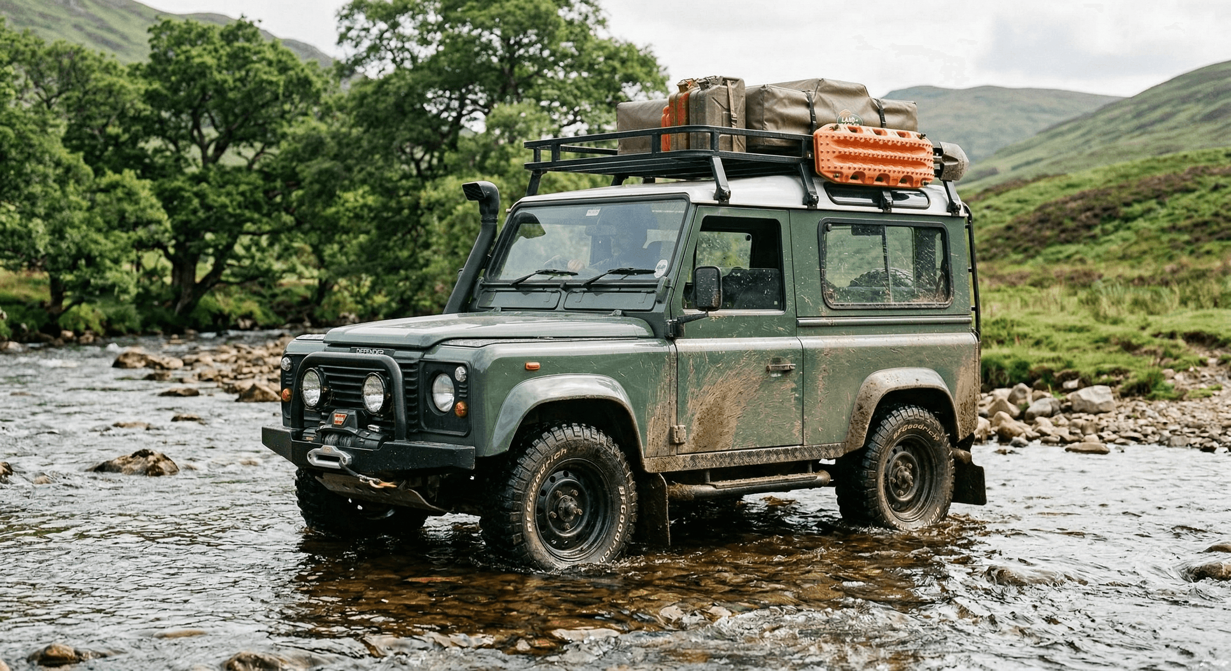 A classic Defender 90 in expedition configuration crossing a river ford, water at axle height, with snorkel fitted and mud caking the aluminum body panels